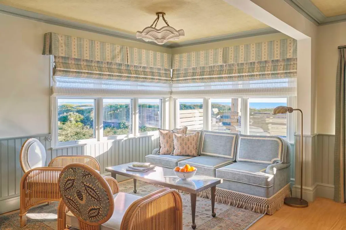 The image showcases a bright, airy studio living area with expansive windows framing stunning sunrise views over the dunes. The inviting space includes a plush sofa adorned with decorative pillows, cozy chairs perfect for relaxing, and a stylish light fixture, all set in a tastefully elegant decor.