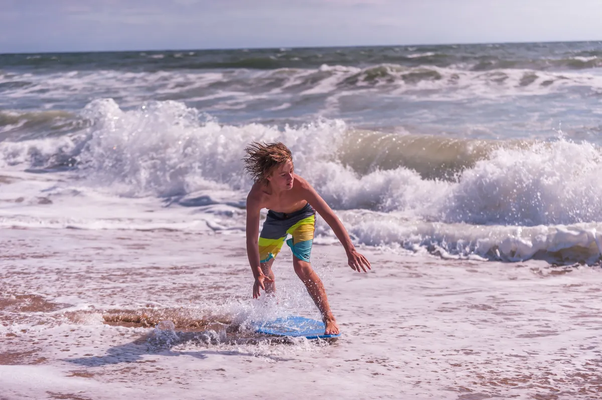 A lively beach scene featuring a boy riding a boogie board amidst the crashing waves, with a sandy shoreline in the background. The image captures the playful spirit and excitement of children enjoying the beach.