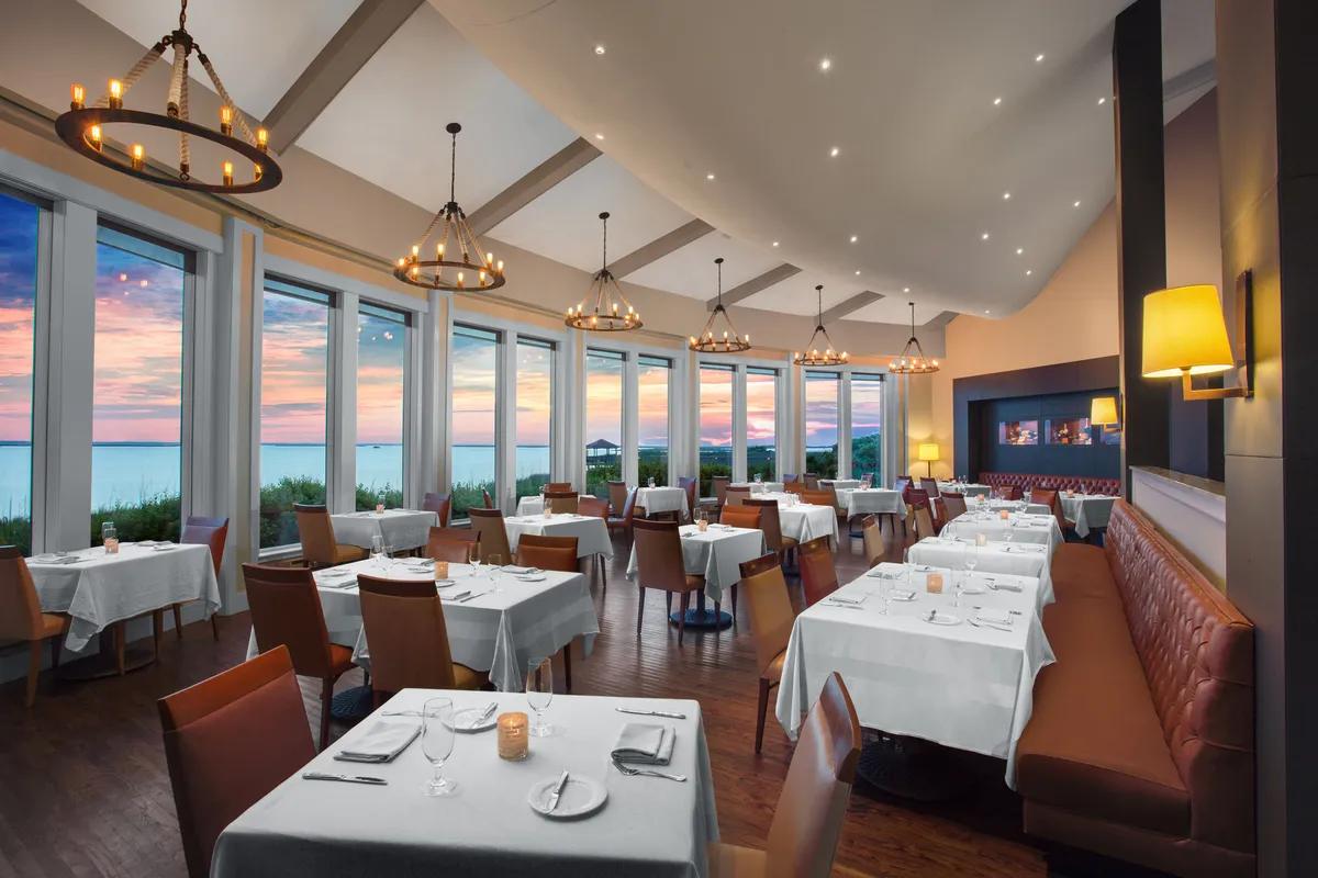 A sophisticated dining area with elegantly set tables, large windows showcasing a vibrant sunset, and chandeliers casting a warm, inviting glow. The scene hints at refined cuisine and elevated dining experiences, with views overlooking the water enhancing the ambiance.