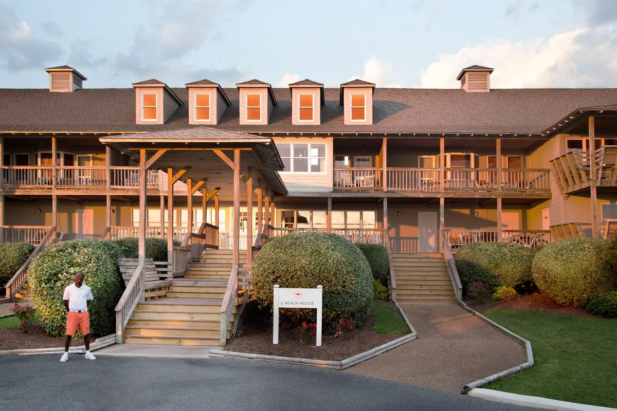 The welcoming main entrance of a beach house, with wooden steps, manicured bushes, and a person enjoying the relaxed ambiance. The sign 'A Beach House' hints at the inviting, home-like feel.