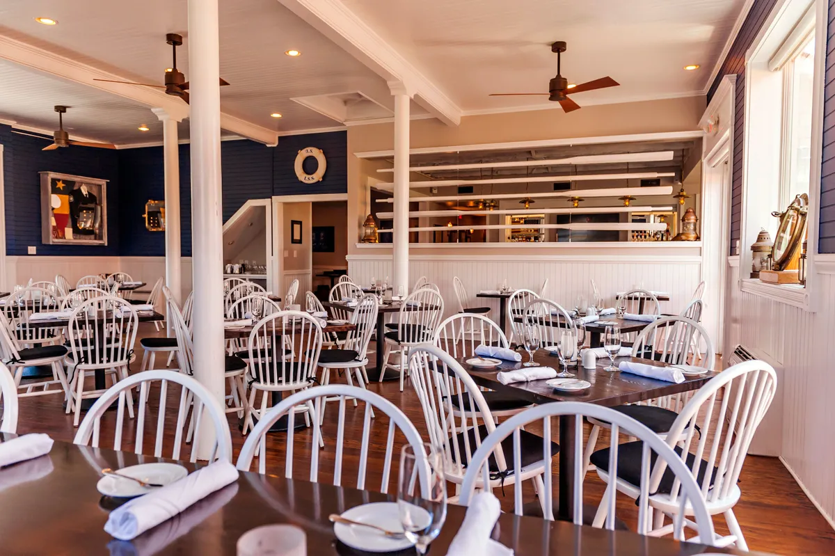 A bright, relaxed dining space with wooden tables, white chairs, cheerful wall art, and a visible bar area. The setup suggests a welcoming environment perfect for families or group celebrations, capturing the comfort and warmth of shared meals.