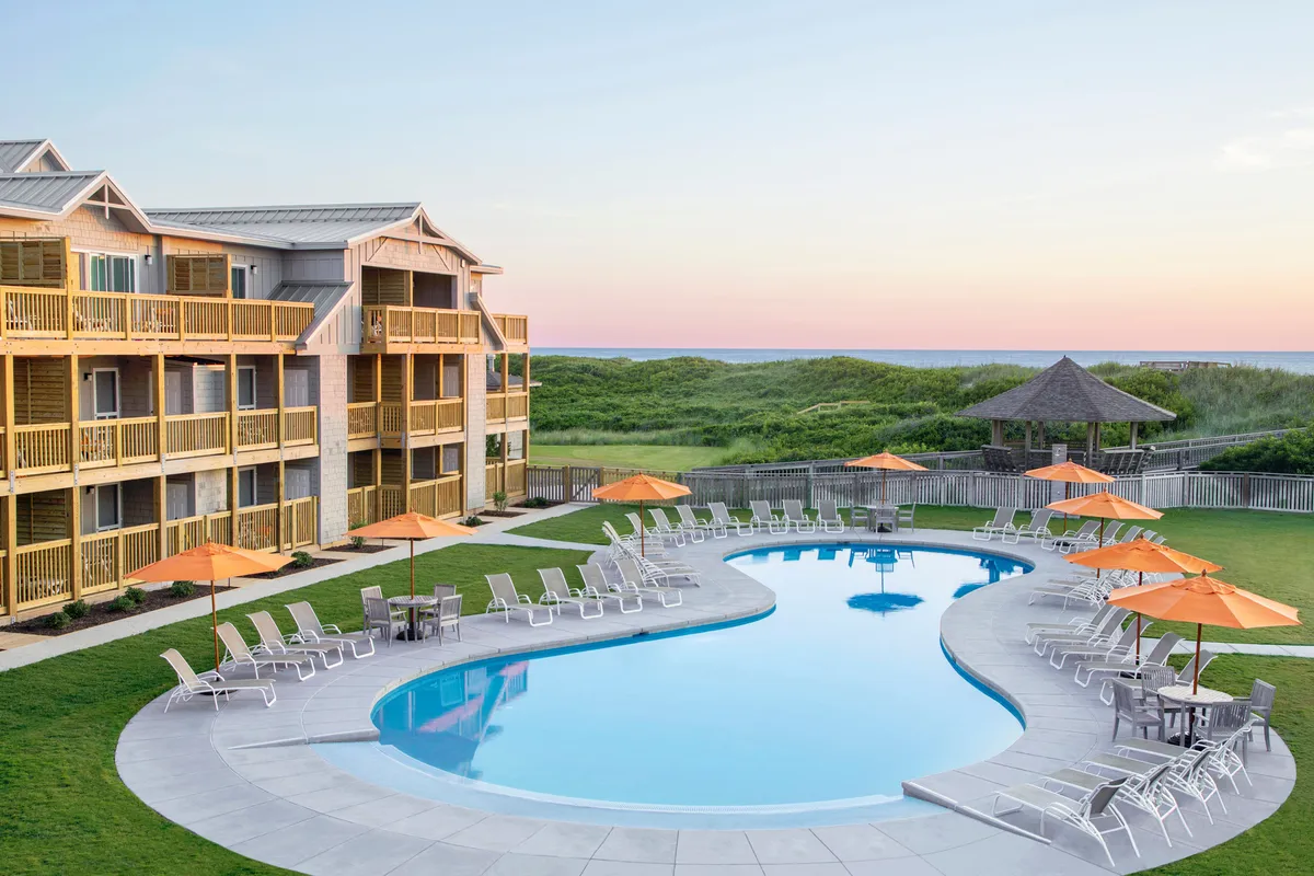 An inviting exterior shot of a serene swimming pool surrounded by outdoor seating, lush gardens, and the resort’s attractive façade. Guests can be seen relaxing and socializing, highlighting the pool as both a place for activity and tranquil leisure.