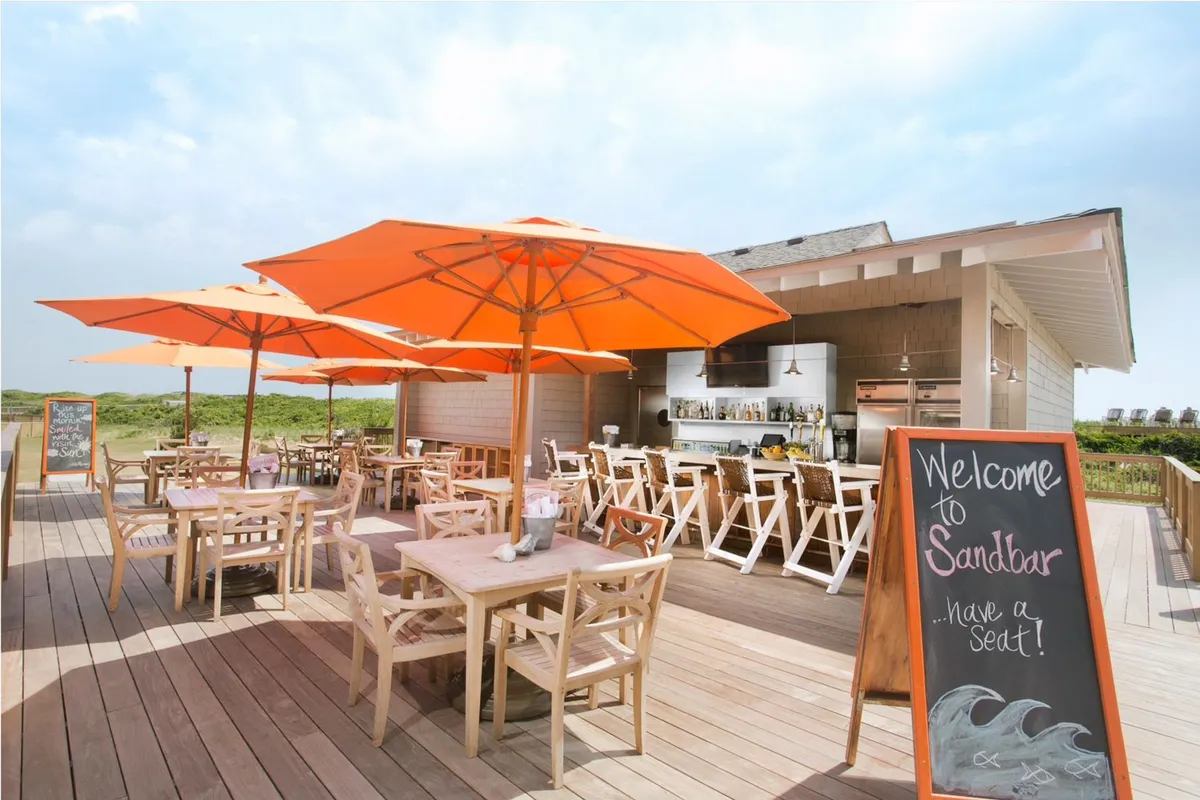 An inviting outdoor dining deck with orange umbrellas, wooden tables and chairs, and a lively bar area overlooks the shoreline. The setting is welcoming and festive, hinting at sunset cocktails and social dining experiences with ocean views.