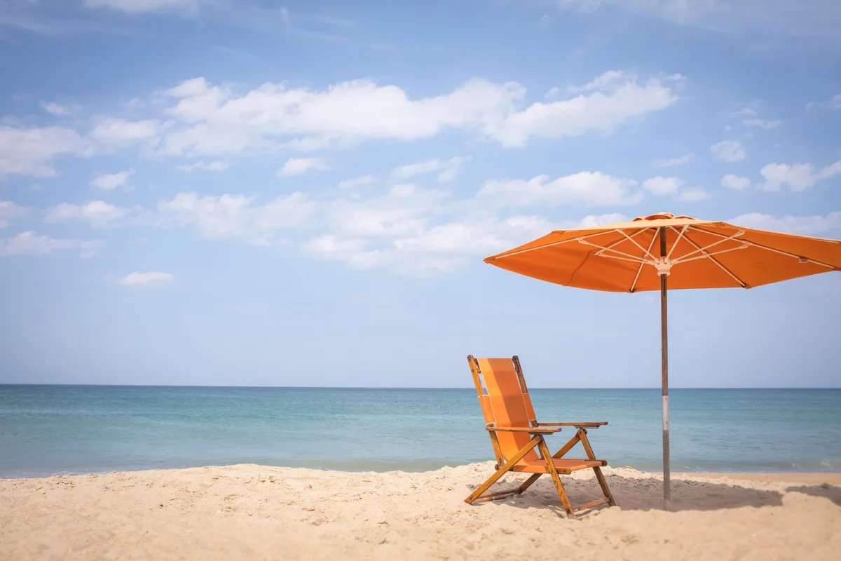 A single beach chair sits under a wide umbrella on soft sand, facing a tranquil ocean. The simple, uncluttered composition evokes peaceful solitude, with gentle waves and an open sky enhancing the sense of escape.