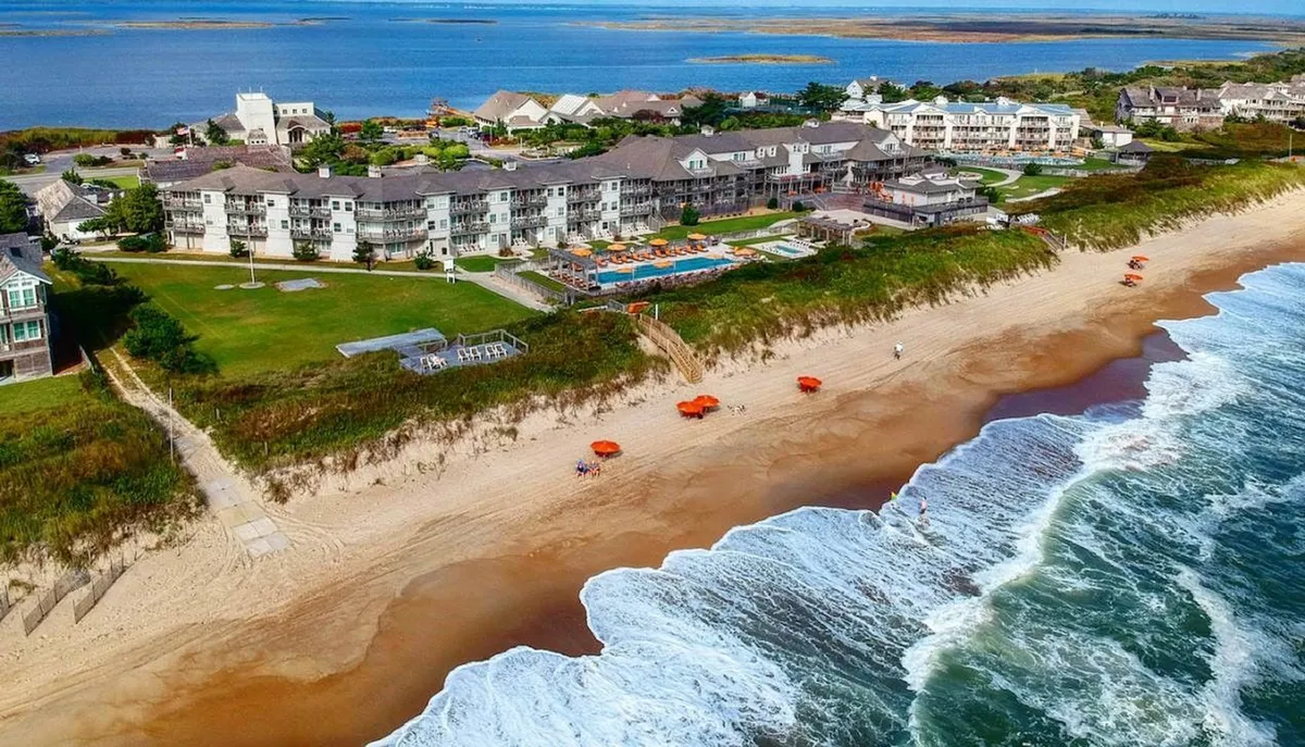 A beachfront pool area merges seamlessly with the sandy shore, featuring hotel buildings, shaded umbrellas, and gentle waves crashing in the background. Families enjoy swimming, sunbathing, and gathering by the water, illustrating the dynamic resort lifestyle.