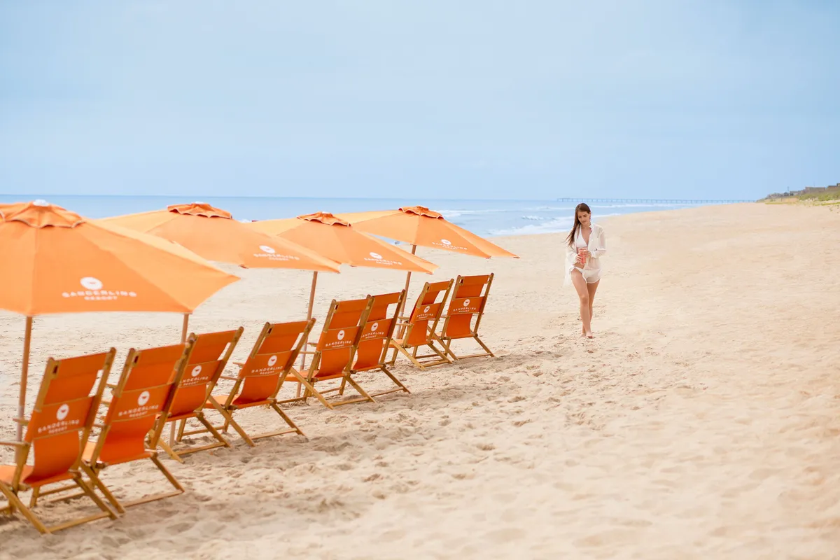A serene stretch of beach is set with inviting chairs and umbrellas, overlooking the sweeping ocean. A woman relaxes under an umbrella, and the expansive shoreline hints at the endless possibilities for adventure and relaxation. The gentle waves and open sky create a tranquil, welcoming scene that captures the Outer Banks' natural beauty.
