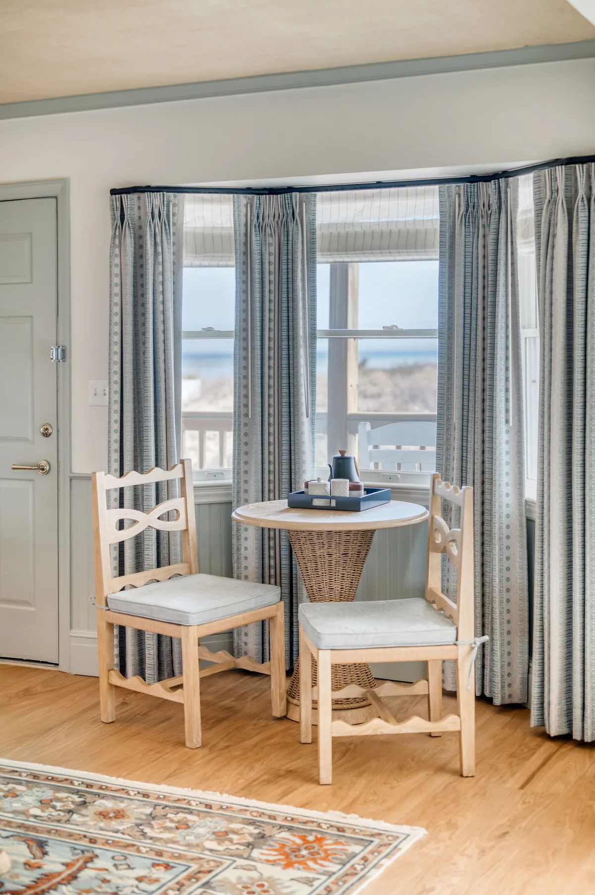 A cozy indoor seating area with a small round table and two wooden chairs is positioned in front of a window, with curtains framing a view of the outdoors. The window reveals a glimpse of coastal scenery, suggesting a tranquil spot for morning coffee or conversation.