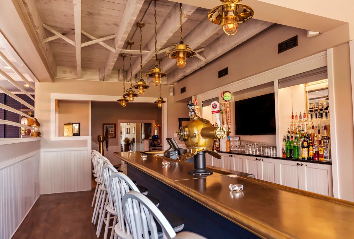 A casual yet stylish bar area featuring a prominent counter, hanging lights, bottles on display, and decorative wall art. The mood is lively but relaxed, suggesting a welcoming spot for drinks and socializing.