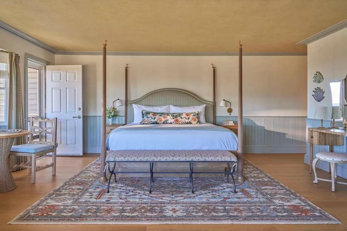 A serene and inviting studio suite with a four-poster king bed dressed in plush linens, an ornate area rug, a cozy small writing desk, and a chair beside the window. Soft daylight filters through, illuminating the peaceful ambiance ideal for restful mornings or relaxing evenings.