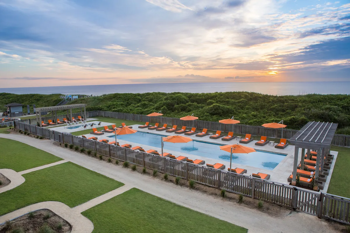 A vibrant outdoor swimming pool with inviting lounge chairs, cheerful orange umbrellas, and stunning ocean views at sunset. Guests are seen swimming, relaxing, and socializing by the poolside, with the tranquil sea providing a breathtaking backdrop.