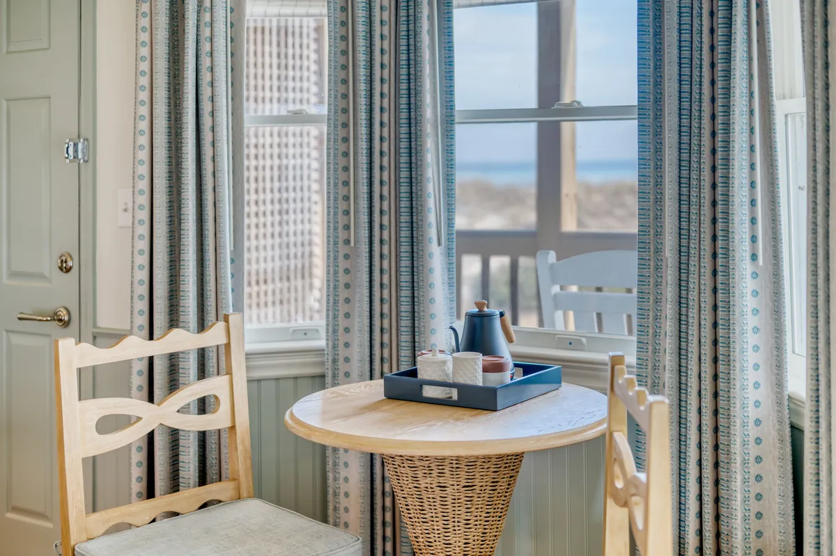 An inviting corner with a wooden table, woven chair, and cups set for coffee, positioned near a window with curtains. The soft lighting and view outside evoke a sense of calm perfect for leisurely mornings.