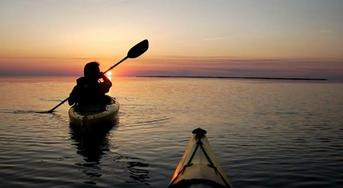 A tranquil scene captures the silhouette of a kayaker gliding across calm water at sunset, with the sky awash in warm hues and the ocean stretching out to the horizon. The peacefulness of the moment highlights the connection to nature and the serene oceanfront setting.